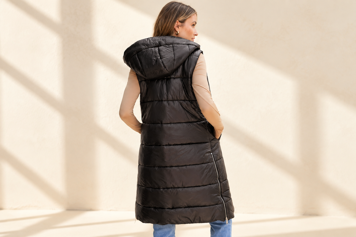 Black hooded long quilted vest coat on a warm ivory boutique background, angle 2